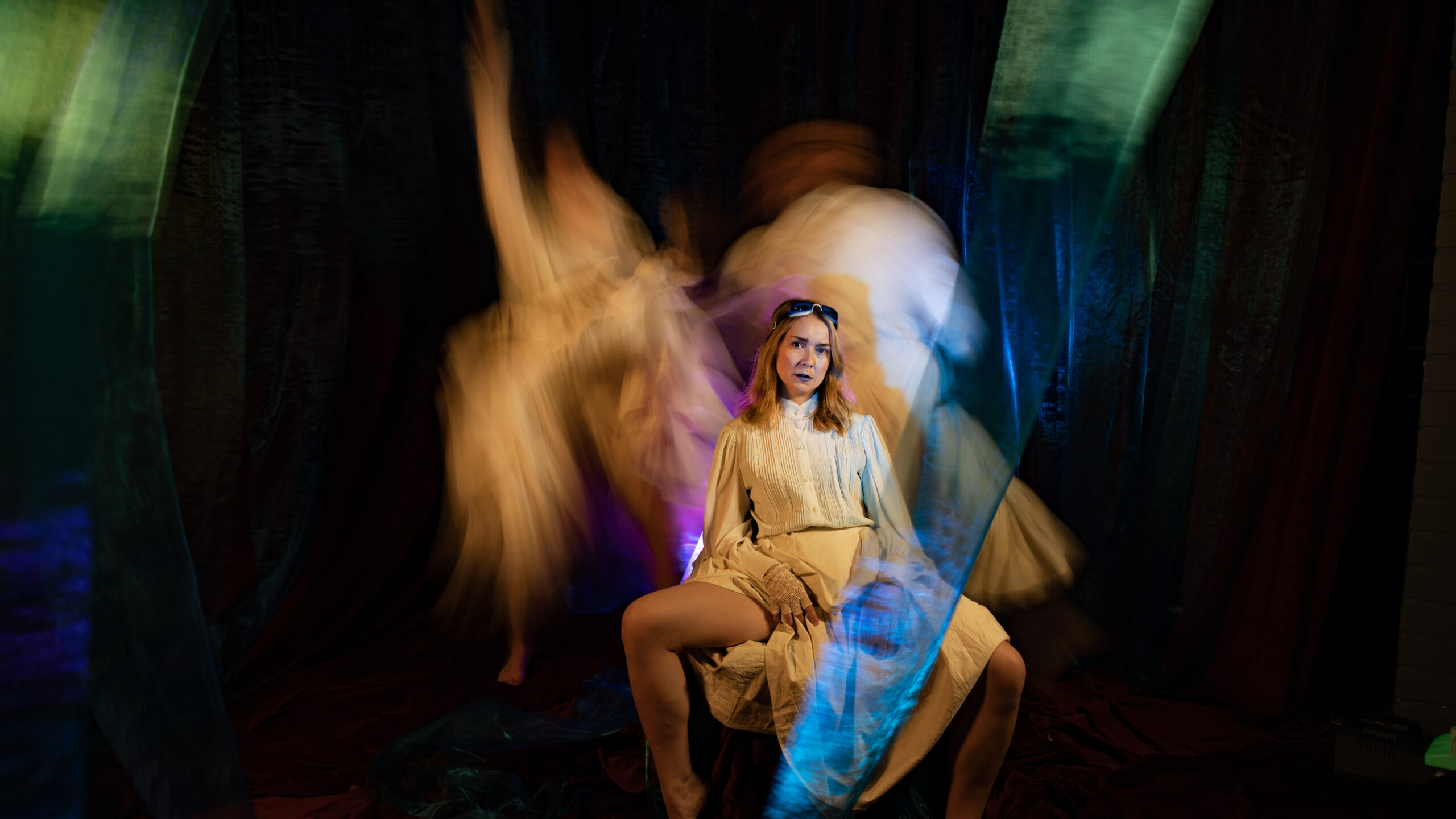 A Person with blonde hair and fair skin in white clothes is sitting legs and looking at camera. Two blurry characters in white clothes at the background.